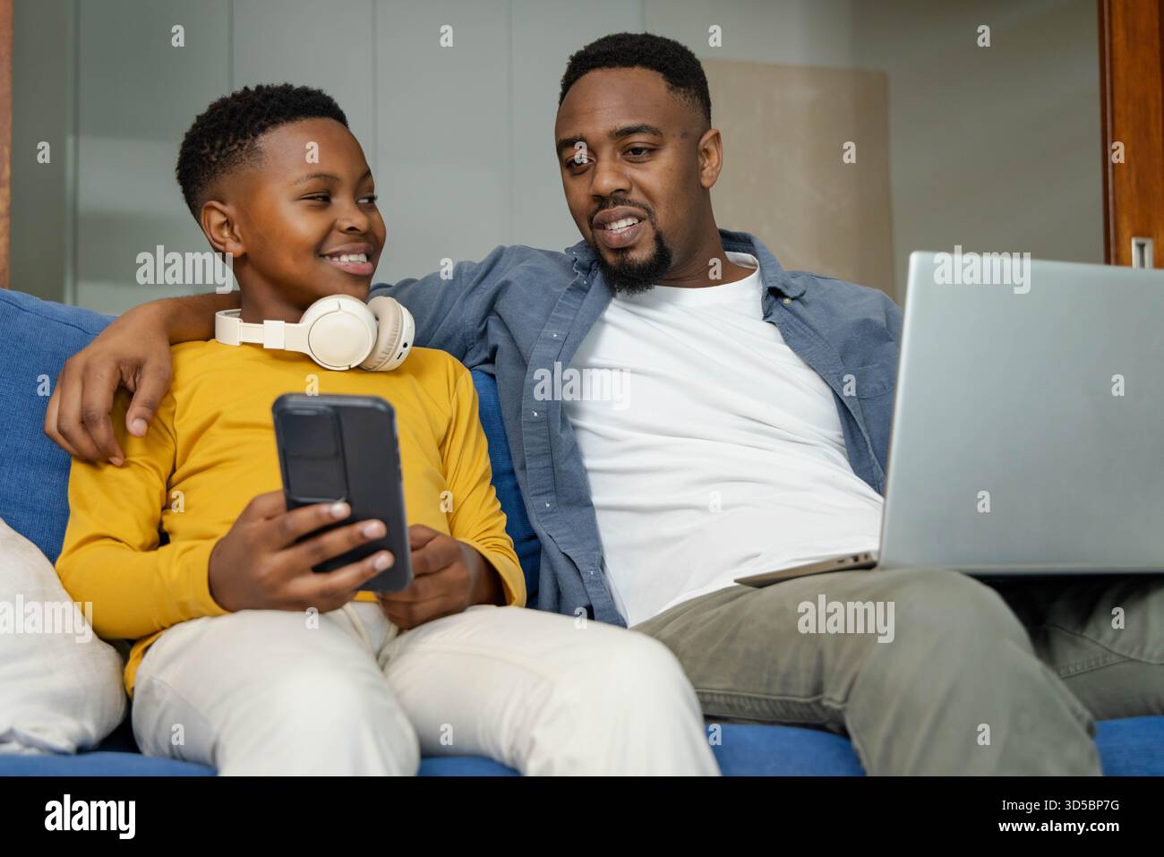Padre e figlio afroamericani che si legano sul divano, utilizzando smartphone e computer portatile insieme Foto Stock