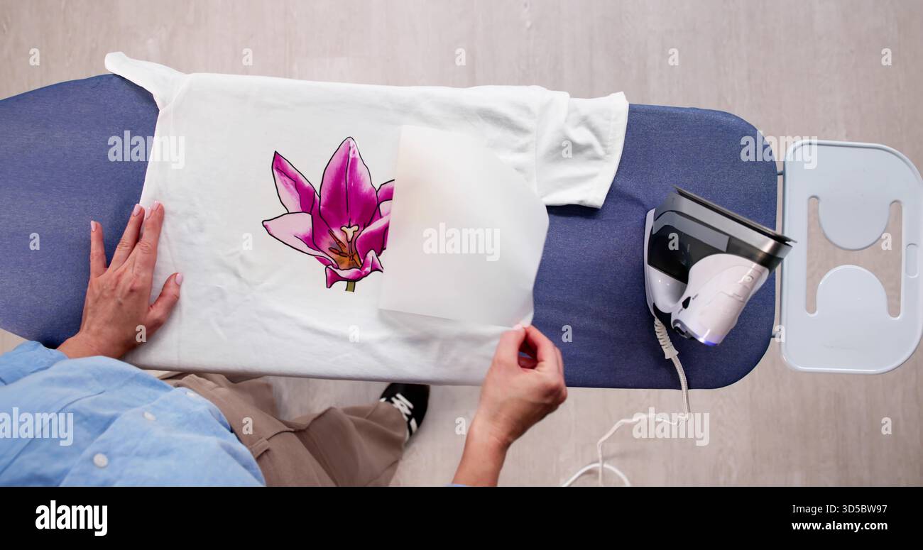 Donna che personalizza il design di una T-shirt con pressa per il trasferimento di ferro a casa. Foto Stock