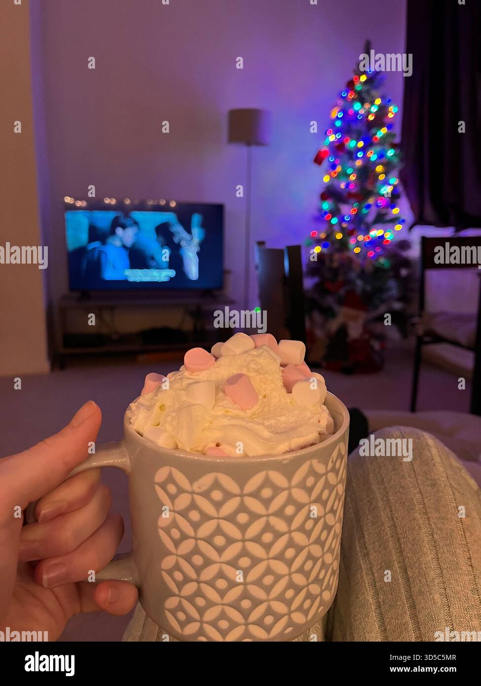 Primo piano di una tazza in ceramica ripiena di cioccolata calda con panna montata e marshmallow, con un albero di Natale decorato sullo sfondo Foto Stock