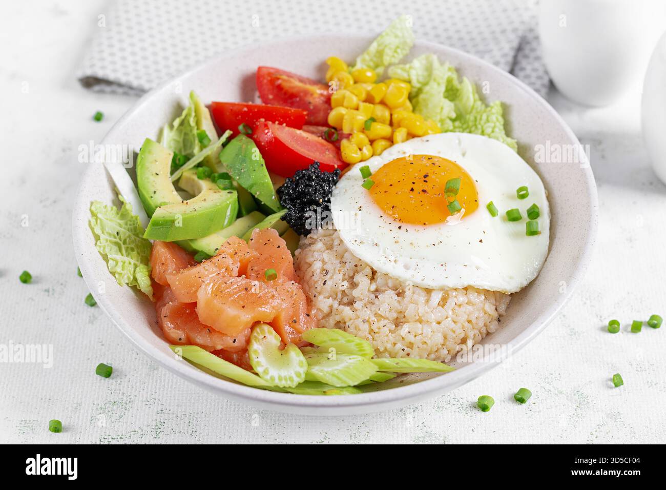 Ciotola con ingredienti freschi come salmone, mais, avocado. pomodori e un uovo con il lato soleggiato su uno sfondo chiaro. Foto Stock