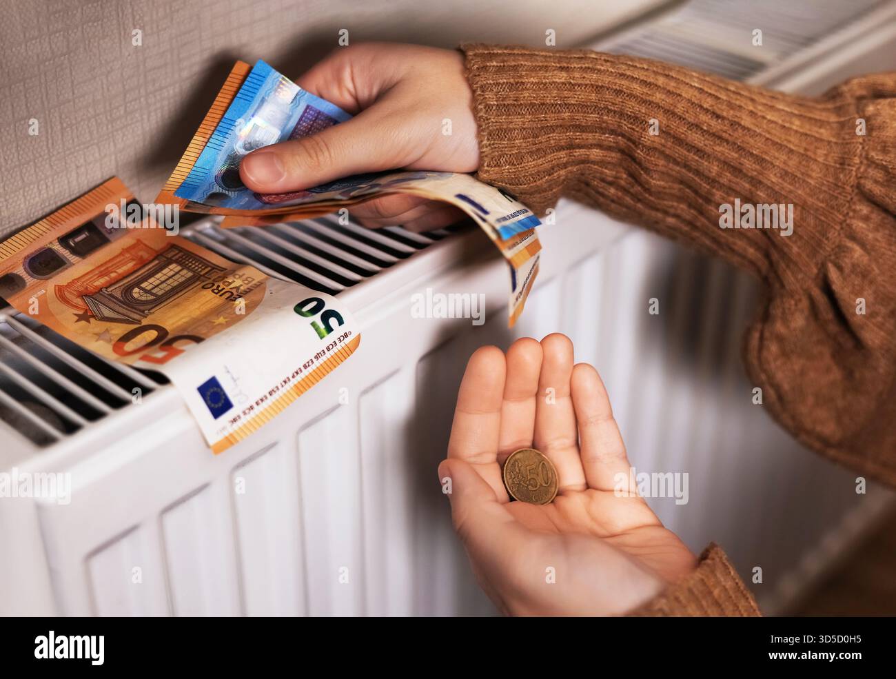 Una ragazza in maglione conta soldi da un radiatore: Banconote da 50 e 20 euro, primo piano di 20 euro e 50 centesimi di euro nelle sue dita. La stanza è fredda, alta Foto Stock