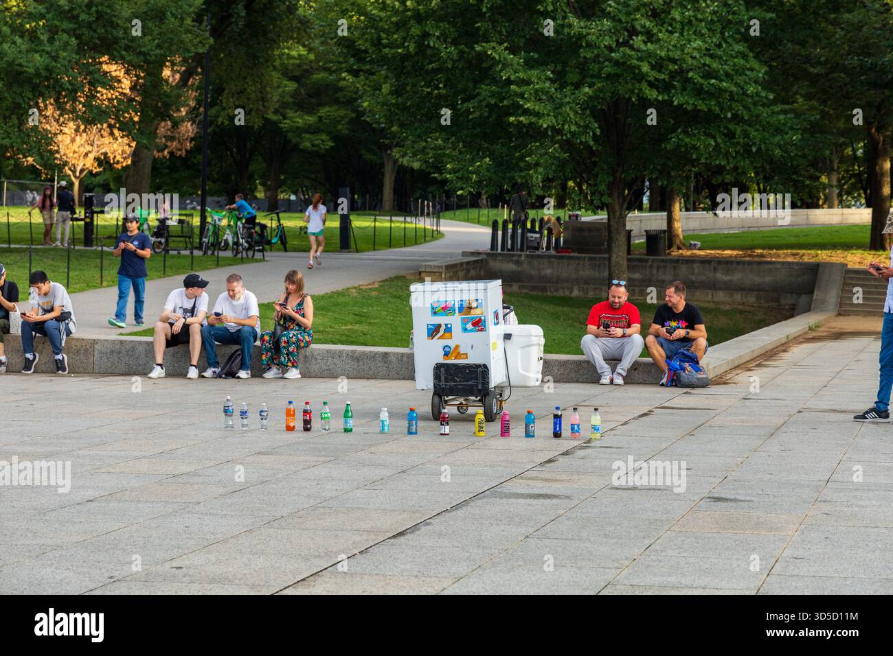 Un gruppo di persone è seduto sul marciapiede in un parco. Un uomo è in piedi davanti a un refrigeratore con un cartello con la scritta "bevande gratuite". CE ne sono diversi Foto Stock