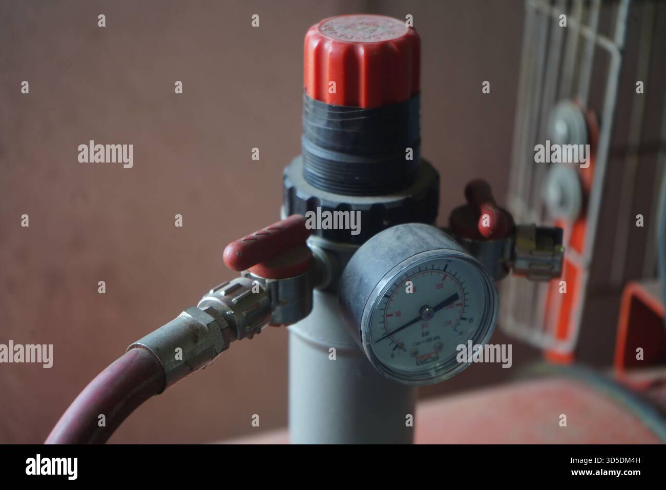 Un regolatore di pressione e un manometro su un compressore d'aria rosso polveroso, incentrato sulla valvola, i raccordi e la strumentazione tecnica in un ambiente di officina. Foto Stock