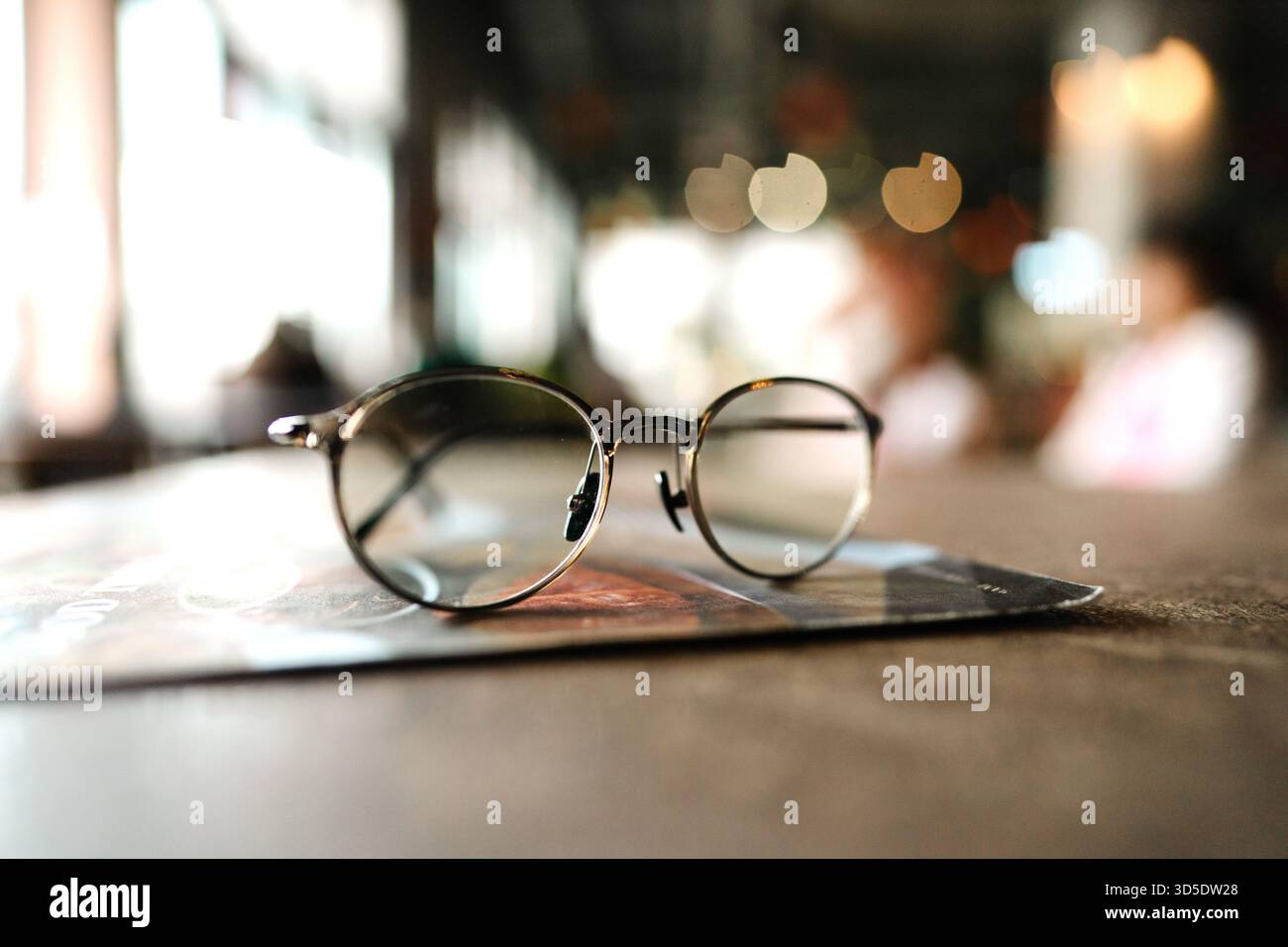 Un paio di occhiali da vista rotondi con montature dorate su una superficie in vetro riflettente, illuminati da luce soffusa, che evocano studio, lettura o atmosfera premurosa Foto Stock