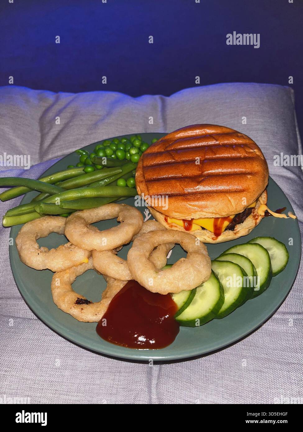 Primo piano di un pasto con hamburger alla griglia con formaggio fuso, anelli di cipolla, piselli, cetrioli e salsa su un piatto. Catturati in interni ad Alicante, Foto Stock