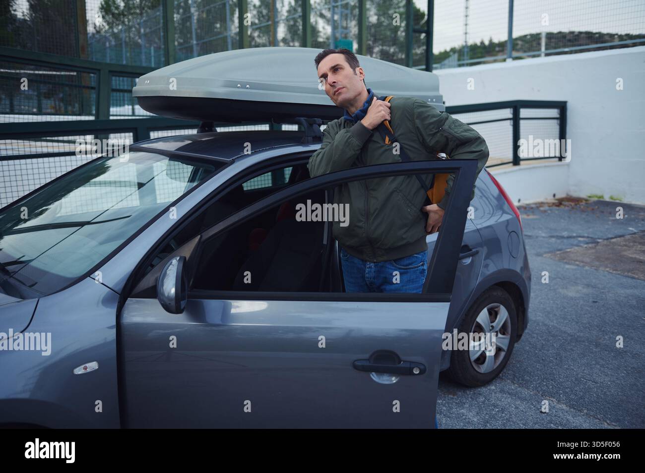 Un uomo si trova accanto a una piccola auto blu con un vano di carico sul tetto, regolando la giacca e preparandosi per un viaggio. La scena suggerisce viaggi, bagagli e jou Foto Stock