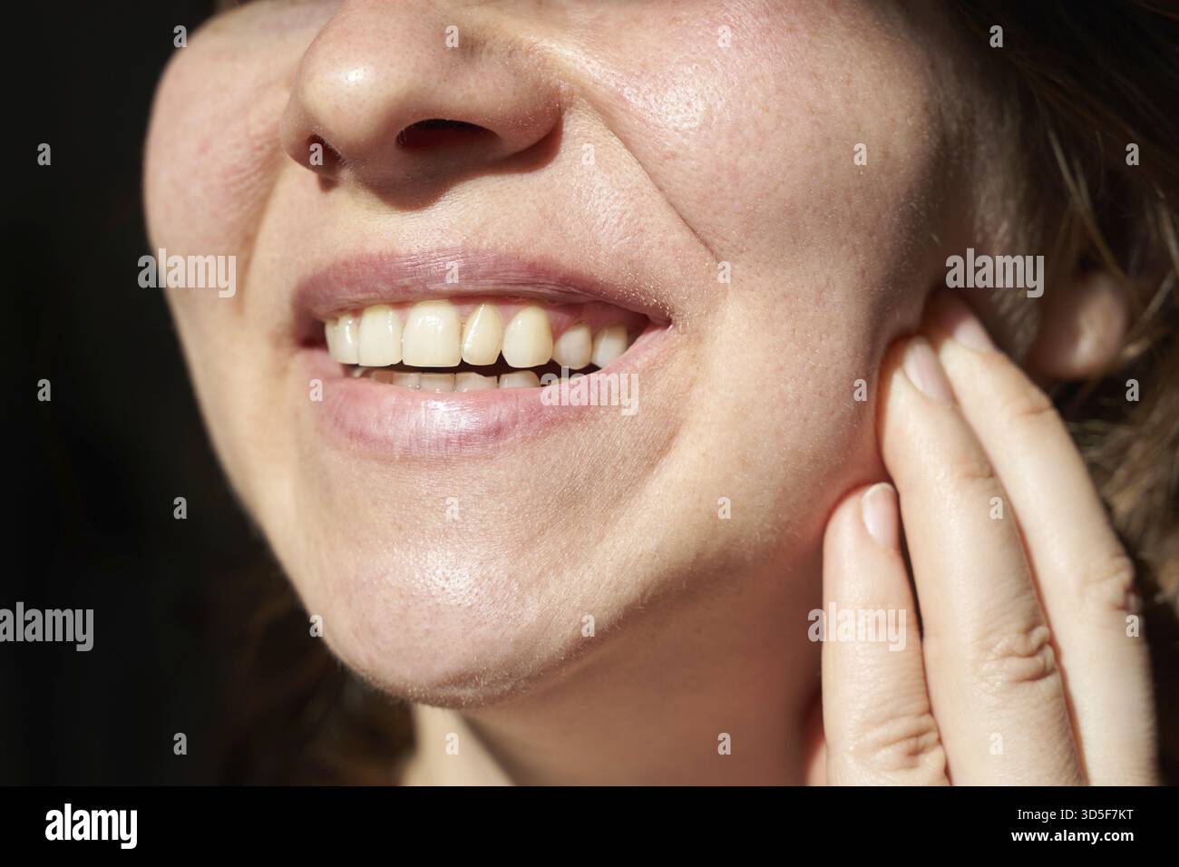 Primo piano di giovane donna caucasica sorridente e toccante guancia con denti sani in piena luce Foto Stock