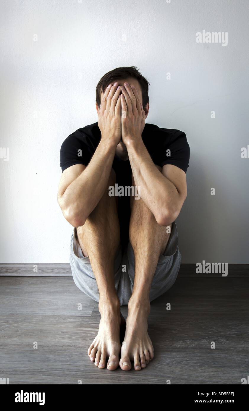 Un uomo seduto sul pavimento contro un muro, che copre il viso con le mani, esprime tristezza, stress o stanchezza emotiva, foto verticale Foto Stock