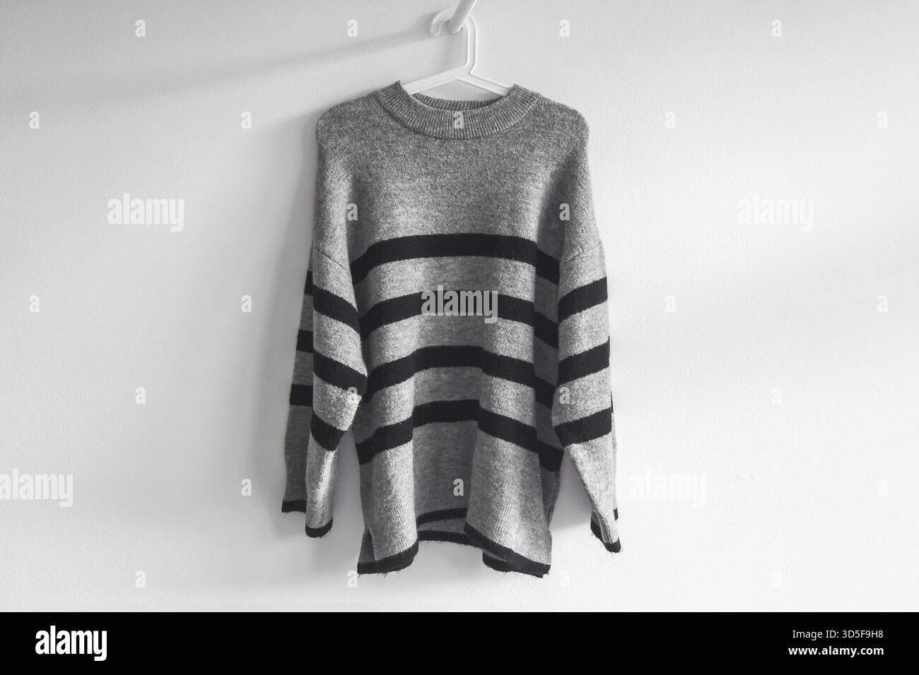 Caldo pullover a righe grigie appeso su un gancio contro una parete bianca Foto Stock