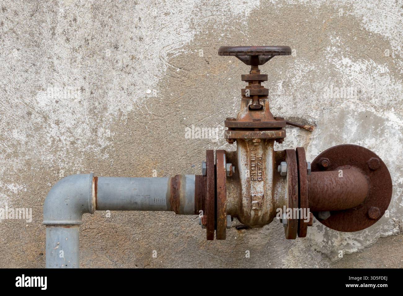 Vecchia valvola industriale arrugginita, all'aperto, con tubi arrugginiti che scompaiono nella parete sullo sfondo. Foto Stock