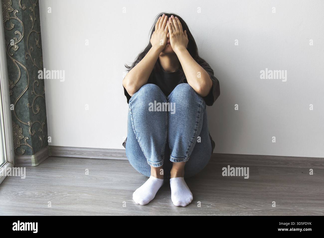 Una giovane donna seduta a terra contro un muro con le ginocchia disegnate, che ricopre il viso con entrambe le mani, esprimendo tristezza, stress o emozione Foto Stock