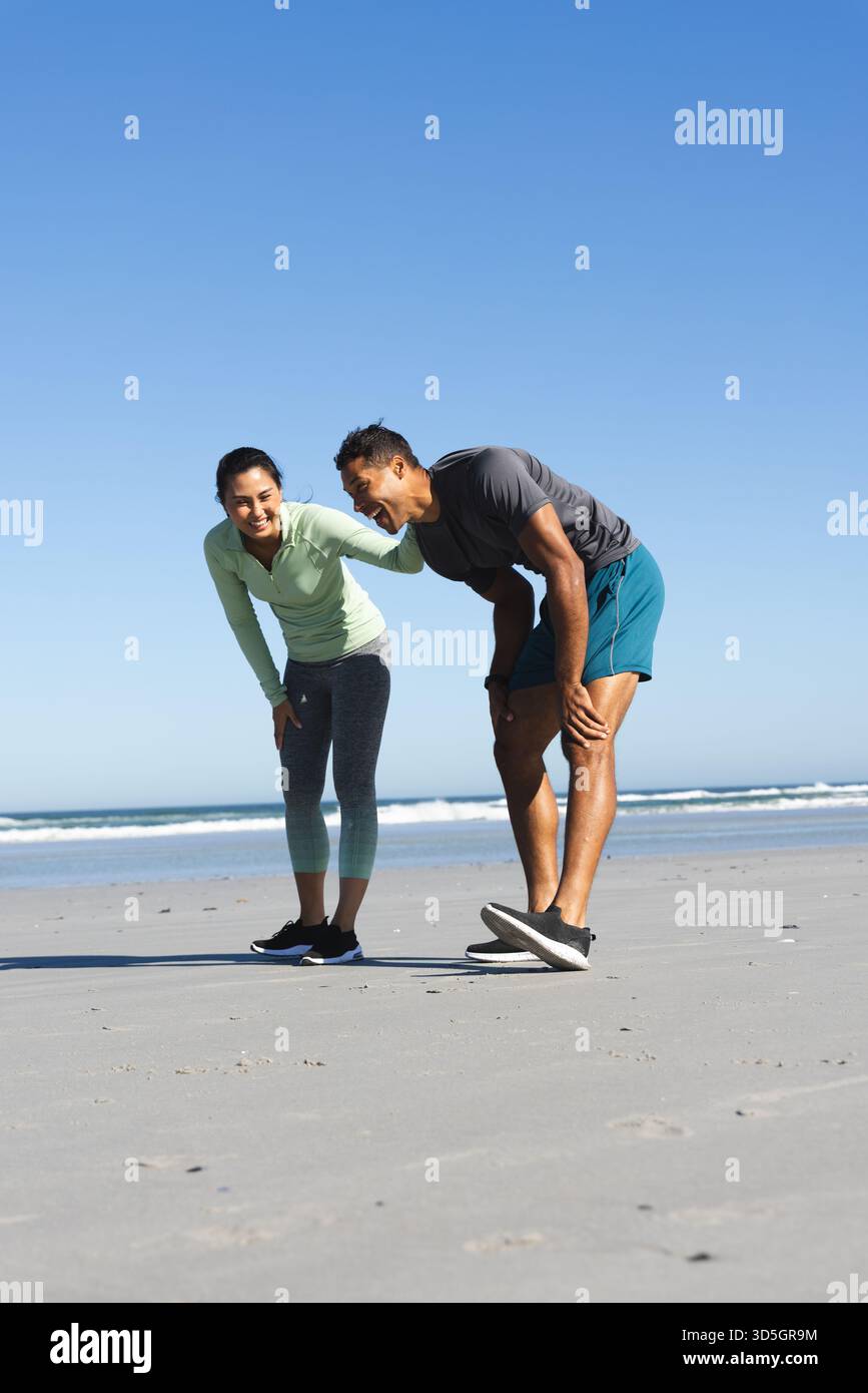 Ridendo e riposando, coppia sulla spiaggia soleggiata dopo aver fatto jogging insieme Foto Stock