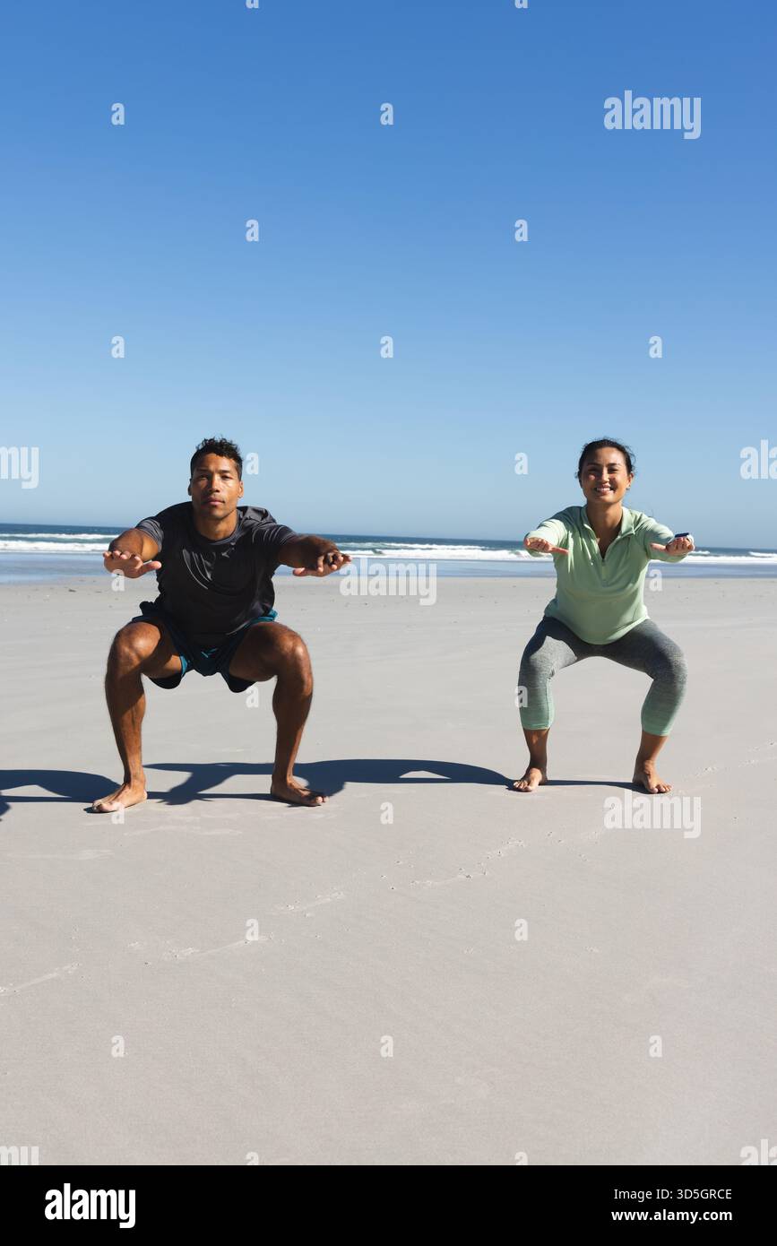 Coppia che si fa esercizio sulla spiaggia, facendo squat insieme sotto il cielo azzurro Foto Stock
