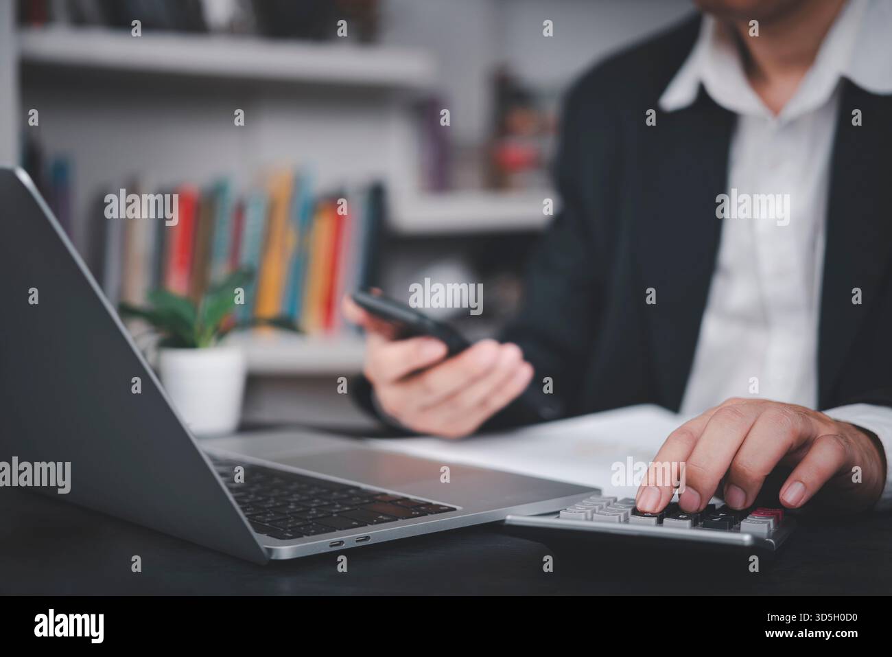 Uomo d'affari che utilizza una calcolatrice e uno smartphone alla scrivania con un computer portatile, simboleggia la pianificazione finanziaria, la contabilità aziendale, l'analisi dei dati, la finanza moderna, b Foto Stock