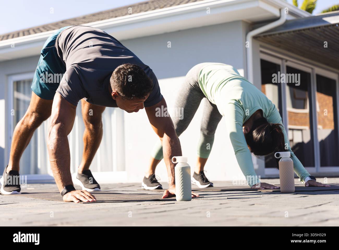 Coppia che pratica yoga all'aperto, mentre si allunga in posa verso il basso con bottiglie d'acqua Foto Stock