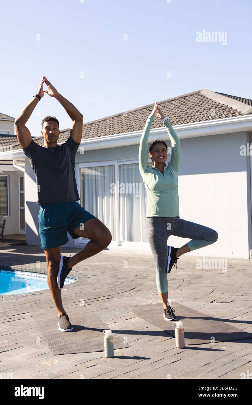 Coppia che pratica yoga all'aperto su tappeti, bilanciandosi in posa sugli alberi a bordo piscina Foto Stock