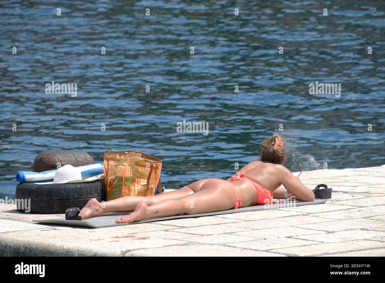 Donna in bikini che fuma sigaretta e guarda l'acqua della baia di Cattaro, Montenegro Foto Stock