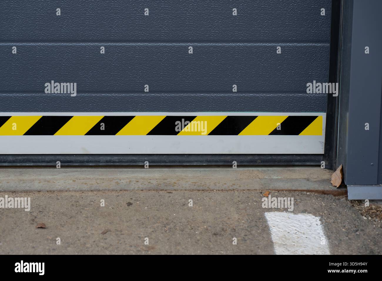 Sezione inferiore della porta di garage industriale con strisce di pericolo gialle nere e superficie del pannello ruvida sopra il grembiule in calcestruzzo, che mostra la posizione dei contrassegni di sicurezza Foto Stock