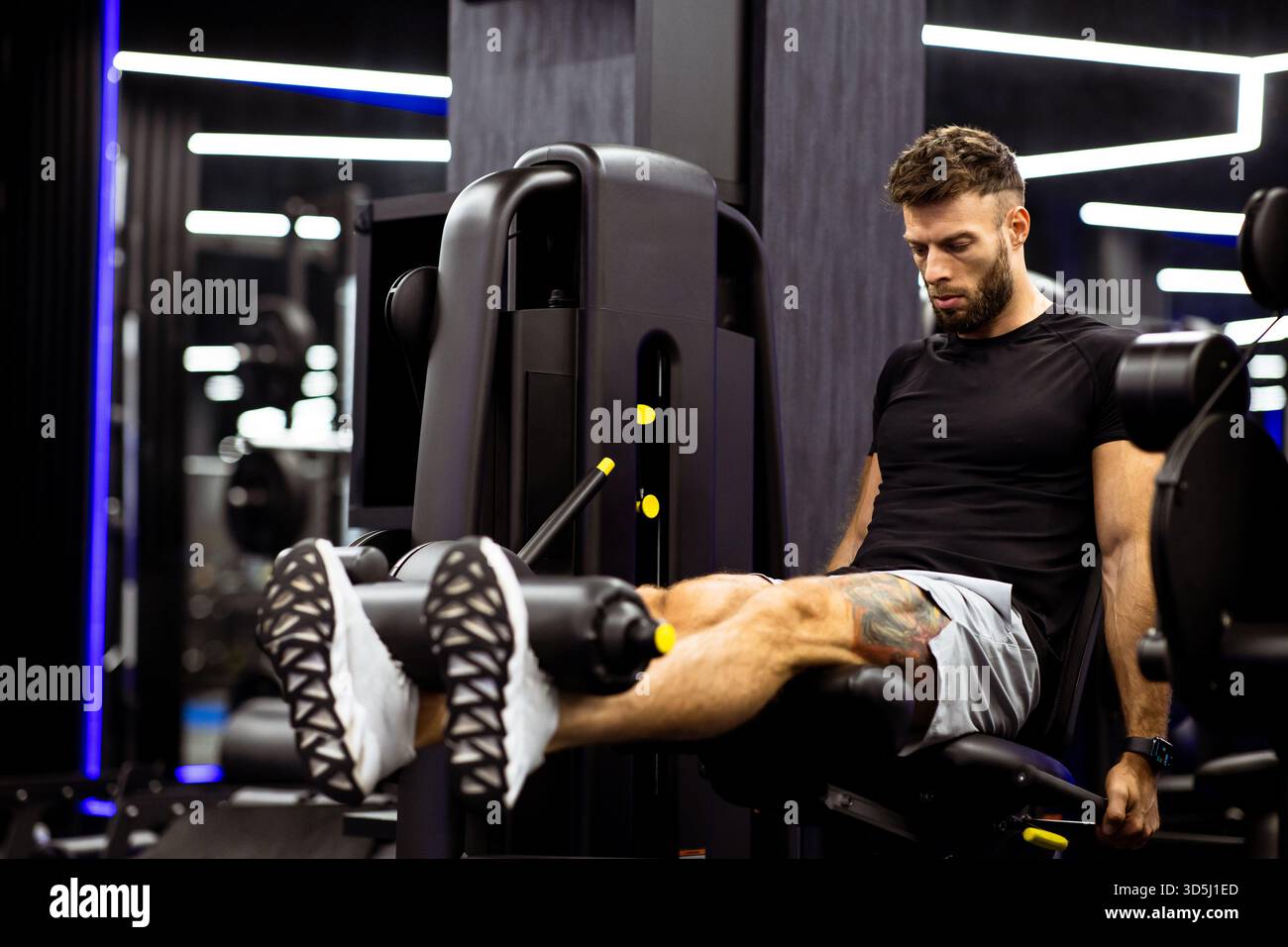 Un uomo fa esercizio su una macchina per allungare le gambe in una palestra moderna. Si concentra sulla costruzione della forza delle gambe. La palestra è ben illuminata con specchi e design elegante, cr Foto Stock
