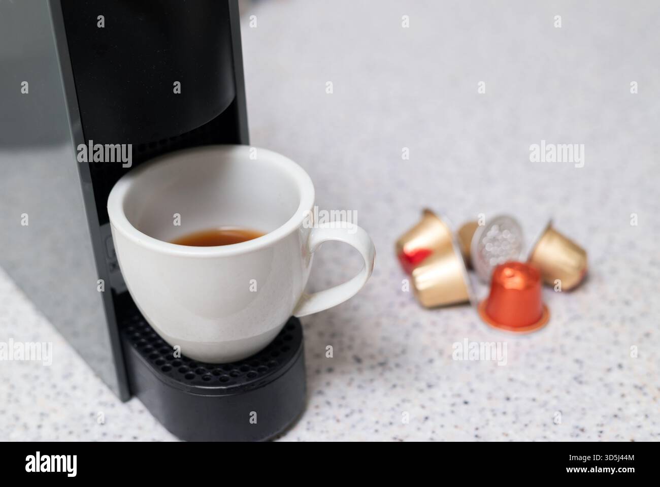 Primo piano di una tazza da caffè bianca con una piccola quantità di caffè, posizionata sotto una macchina a capsule nera con cialde colorate nelle vicinanze Foto Stock