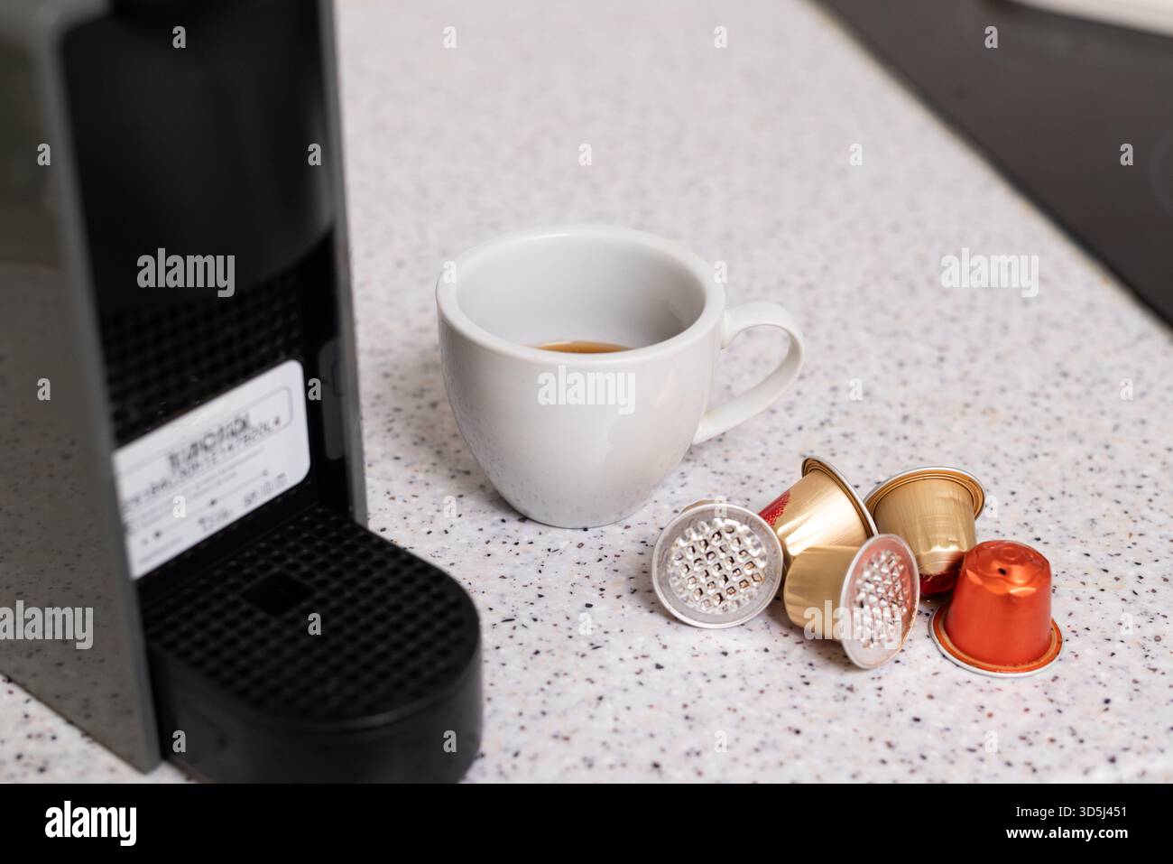 Primo piano di una tazza da caffè bianca con una piccola quantità di caffè, posizionata sotto una macchina a capsule nera con cialde colorate nelle vicinanze Foto Stock