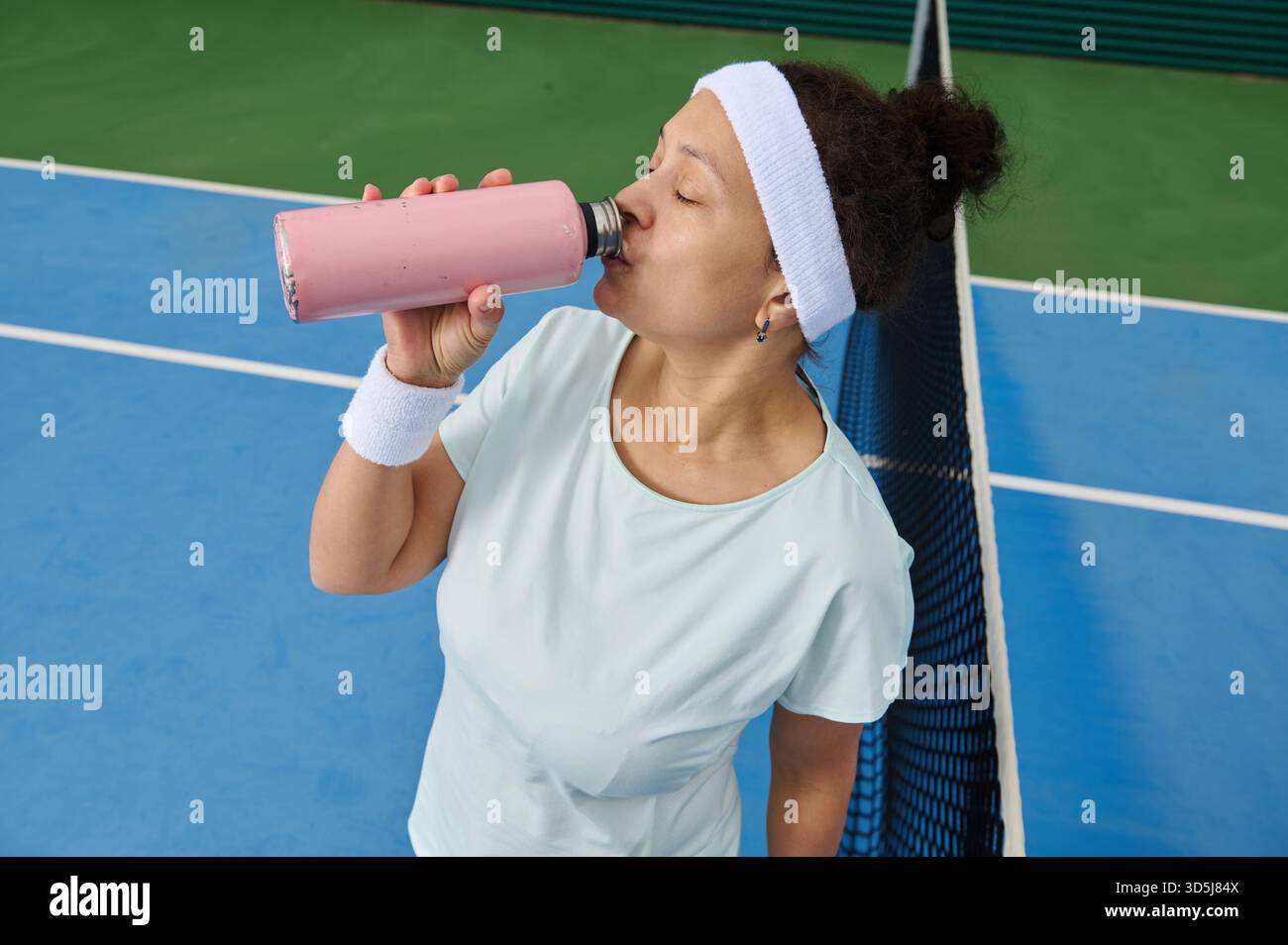 Una donna atletica si prende una pausa su un campo da tennis, sorseggiando un frullato rosa. Indossa un cerchietto bianco e un braccialetto, appoggiandosi casualmente alla rete W Foto Stock