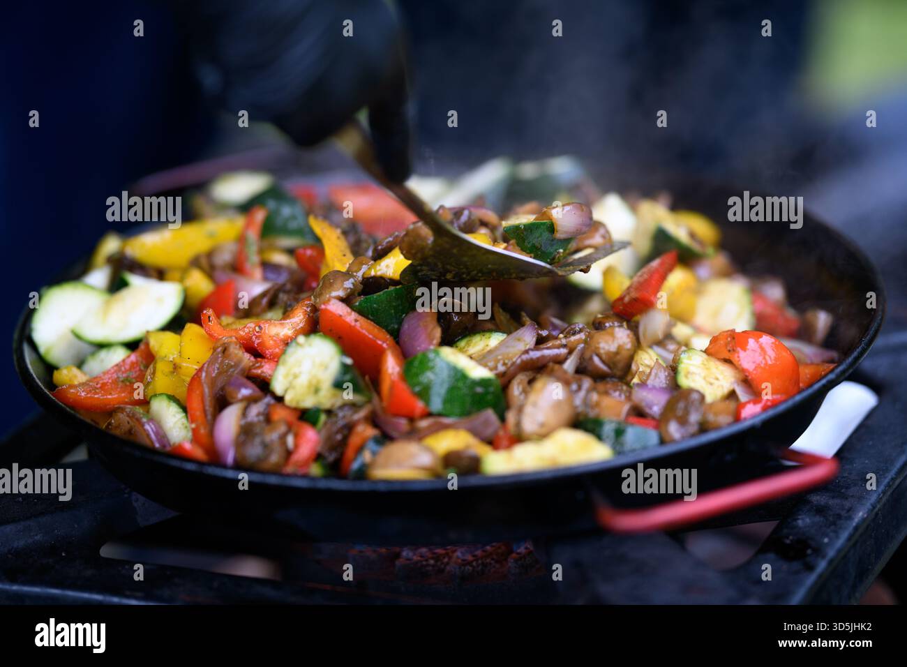 Primo piano di uno chef in un giardino con guanti neri in nitrile che cucinano verdure fresche assortite in una padella su un piano cottura a gas Foto Stock