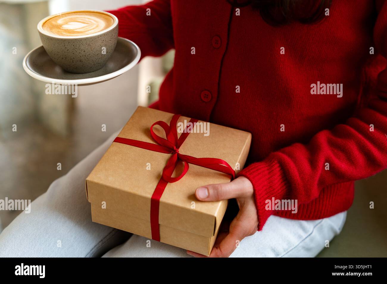Donna in maglione rosso che tiene in mano una tazza di caffè e una confezione regalo con nastro rosso â€” accogliente atmosfera natalizia Foto Stock