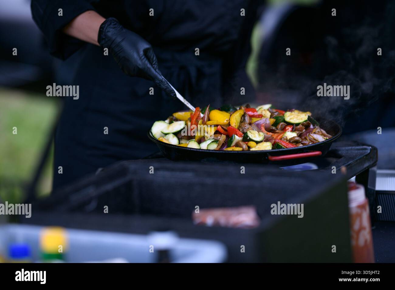 Primo piano di uno chef in un giardino con guanti neri in nitrile che cucinano verdure fresche assortite in una padella su un piano cottura a gas Foto Stock