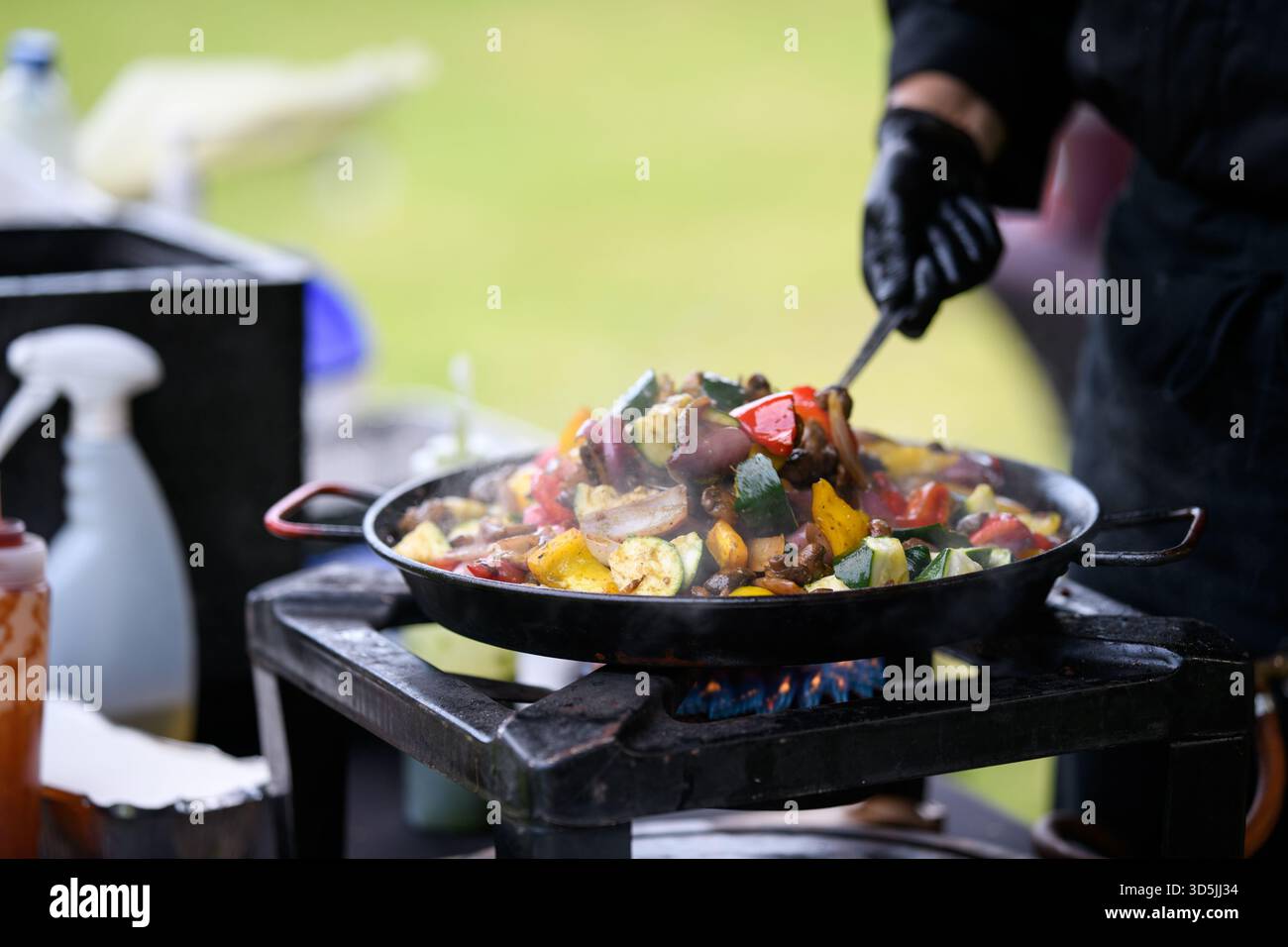 Primo piano di uno chef in un giardino con guanti neri in nitrile che cucinano verdure fresche assortite in una padella su un piano cottura a gas Foto Stock