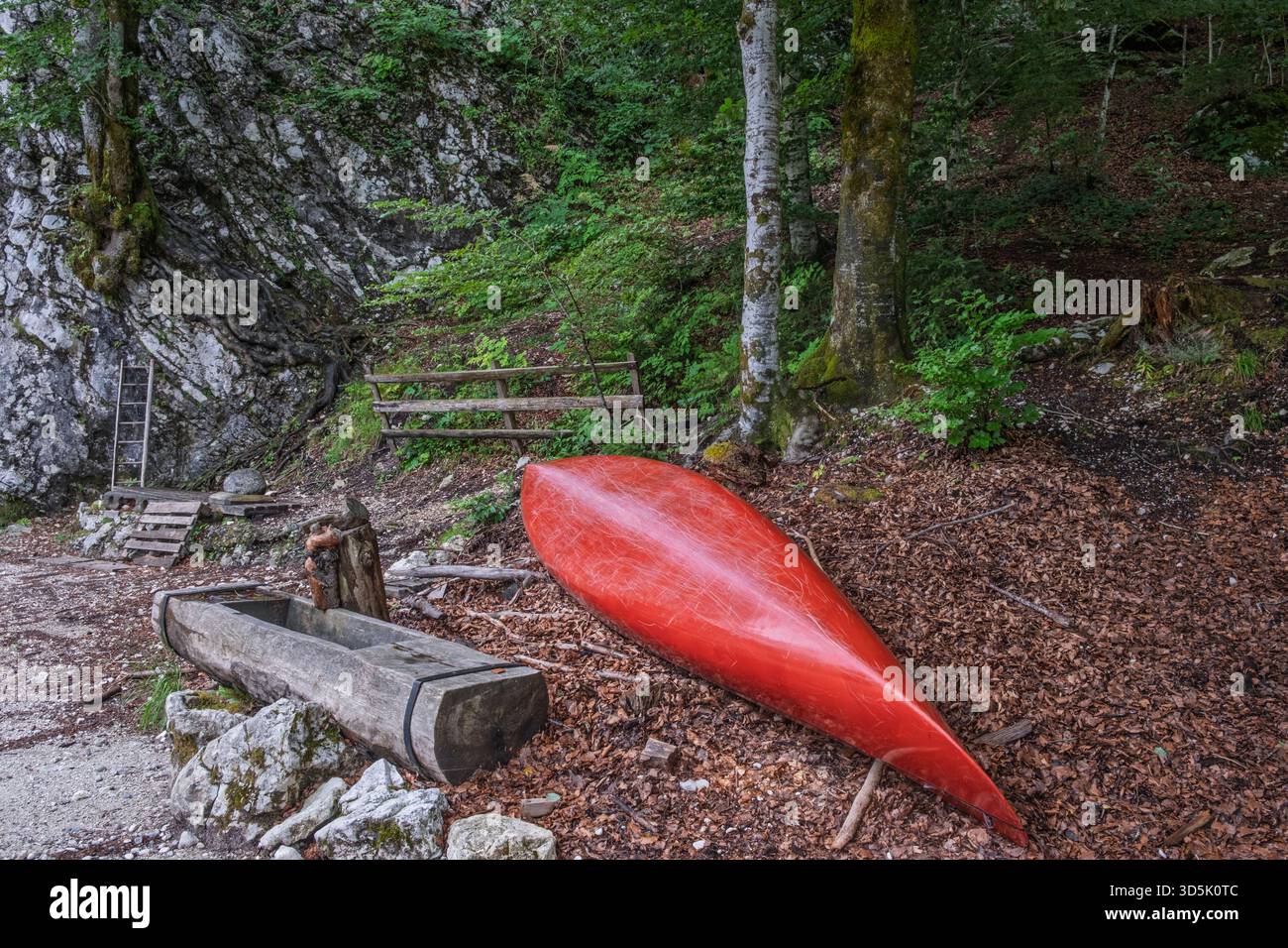 Canoa rossa che riposa a testa in giù in una foresta che si radura accanto a rustiche strutture in legno e terreno roccioso, tranquilla scena naturale all'aperto. Foto Stock