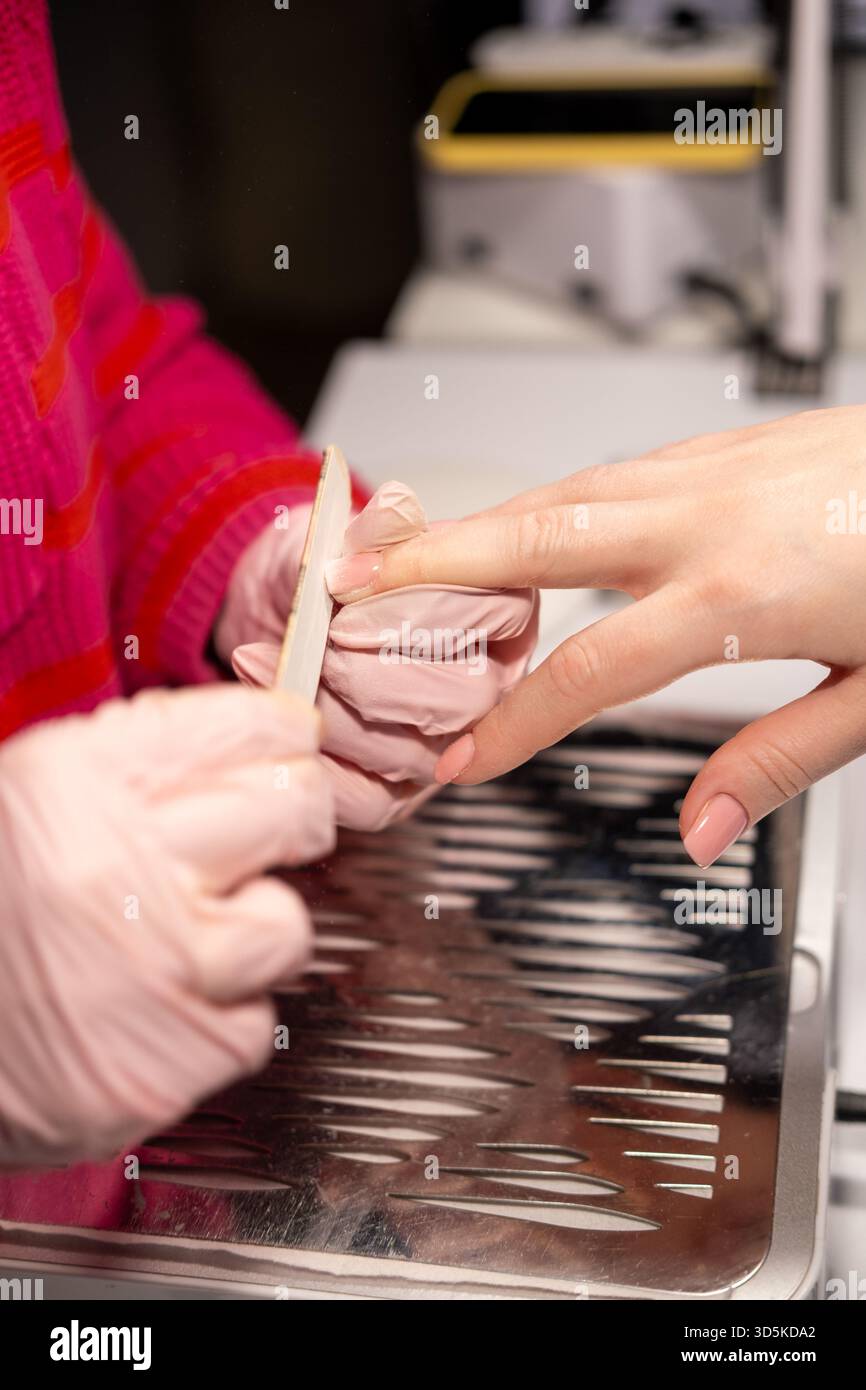Procedura di manicure. Lima per unghie. Manicurista professionista donna con le mani in guanti protettivi rosa che utilizzano il bordo di smeriglio nel salone di bellezza tenendo il dito dei clienti. Foto Stock