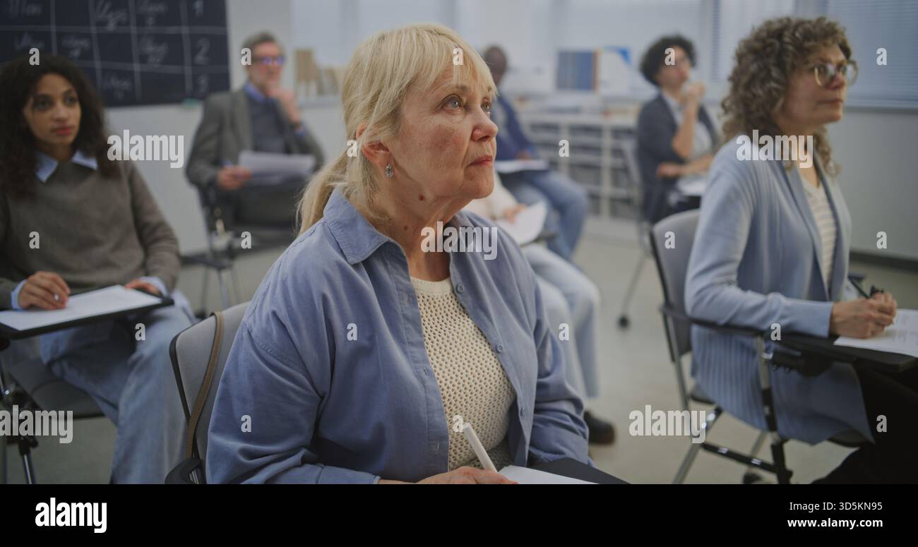 Gli studenti adulti siedono sulla scrivania, concentrandosi sulle attività scritte mentre ascoltano l'istruttore. I diversi studenti rimangono coinvolti, prendendo appunti e completando le attività in un ambiente tranquillo e strutturato in classe. Foto Stock