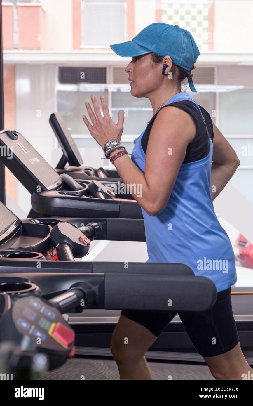 Una donna di mezza età che si allena e corre sul tapis roulant, indossando un berretto e un abbigliamento sportivo blu. Foto Stock