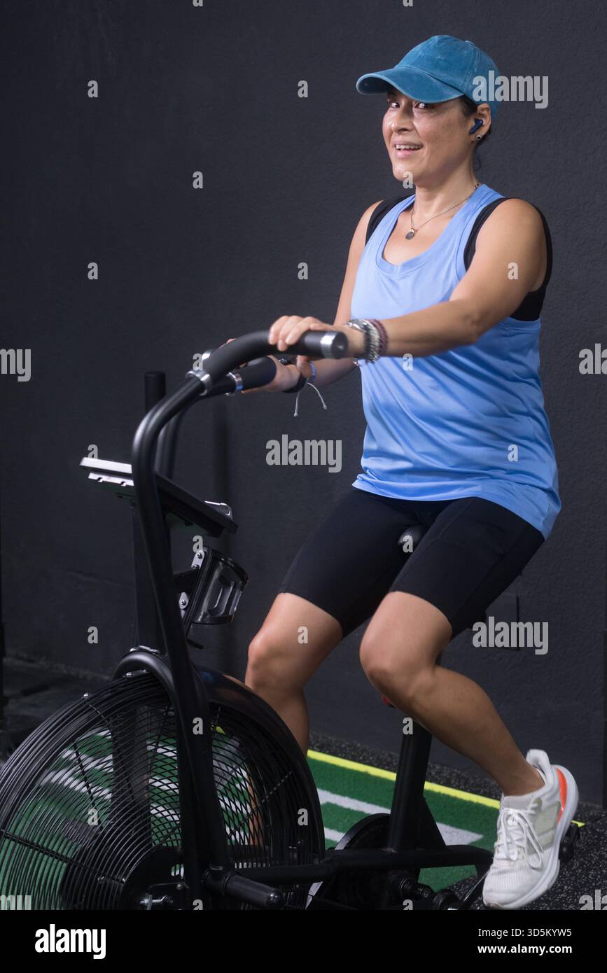 Una donna di mezza età che si allena su una macchina Air bike, indossa un berretto e un abbigliamento sportivo blu. Foto Stock