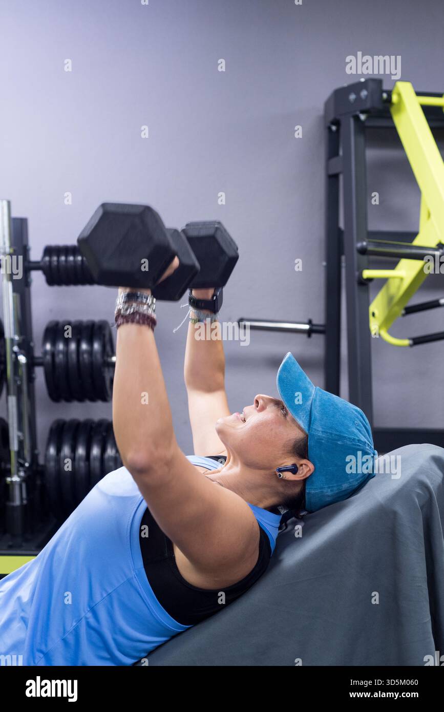 Donna di mezza età che si allena sollevando i manubri su una panca inclinata, indossando un berretto e un abbigliamento sportivo blu. Foto Stock