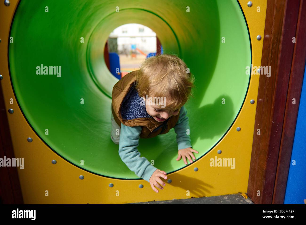 Un bambino esplora un tunnel verde luminoso in un parco giochi, indossando un gilet marrone e maniche blu, assaporando una divertente giornata autunnale. Cattura il chil. Senza pensieri Foto Stock