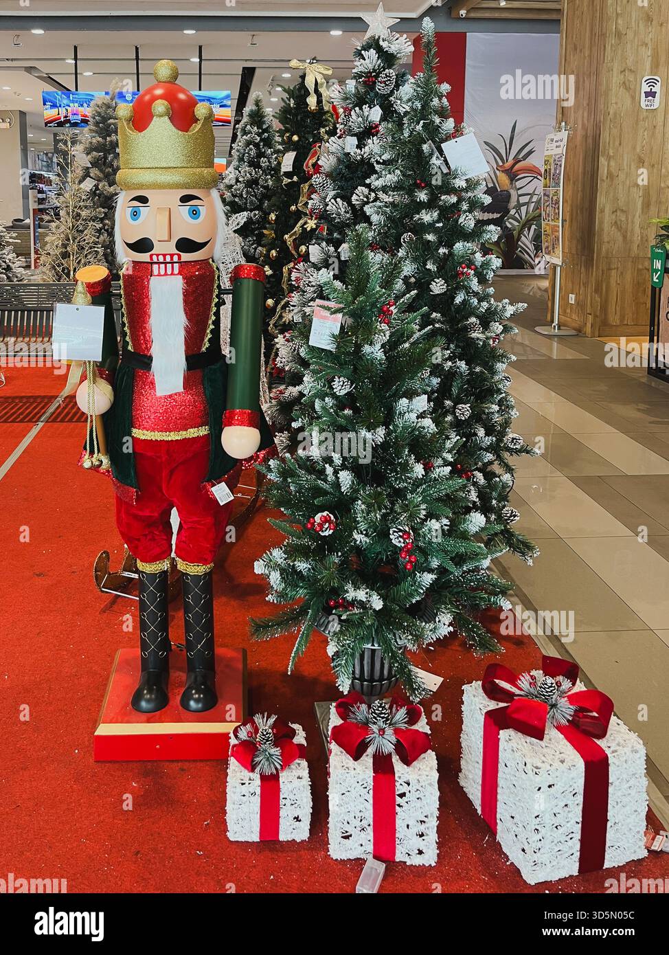 Balikpapan, Indonesia - 16 novembre 2025. Una grande statua dello Schiaccianoci e alberi di Natale artificiali, circondati da scatole regalo decorative, come Foto Stock