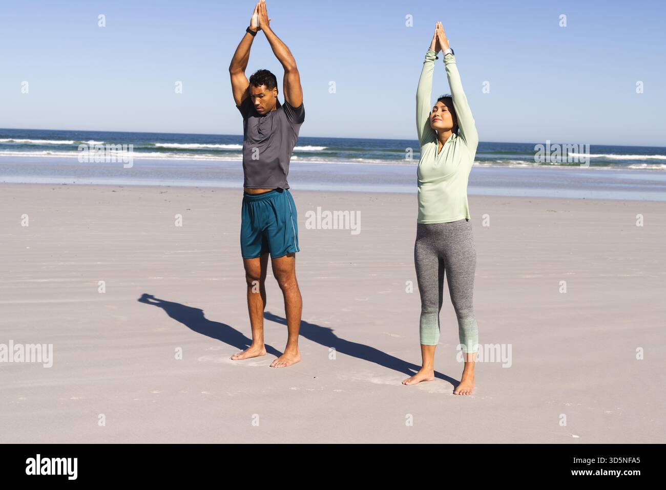 Coppia che pratica yoga sulla spiaggia, allungando le braccia verso l'alto sotto il cielo limpido Foto Stock