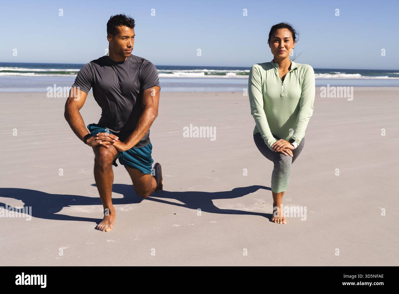 Coppia che si fa esercizio sulla spiaggia, facendo affondamenti insieme sotto il cielo azzurro Foto Stock
