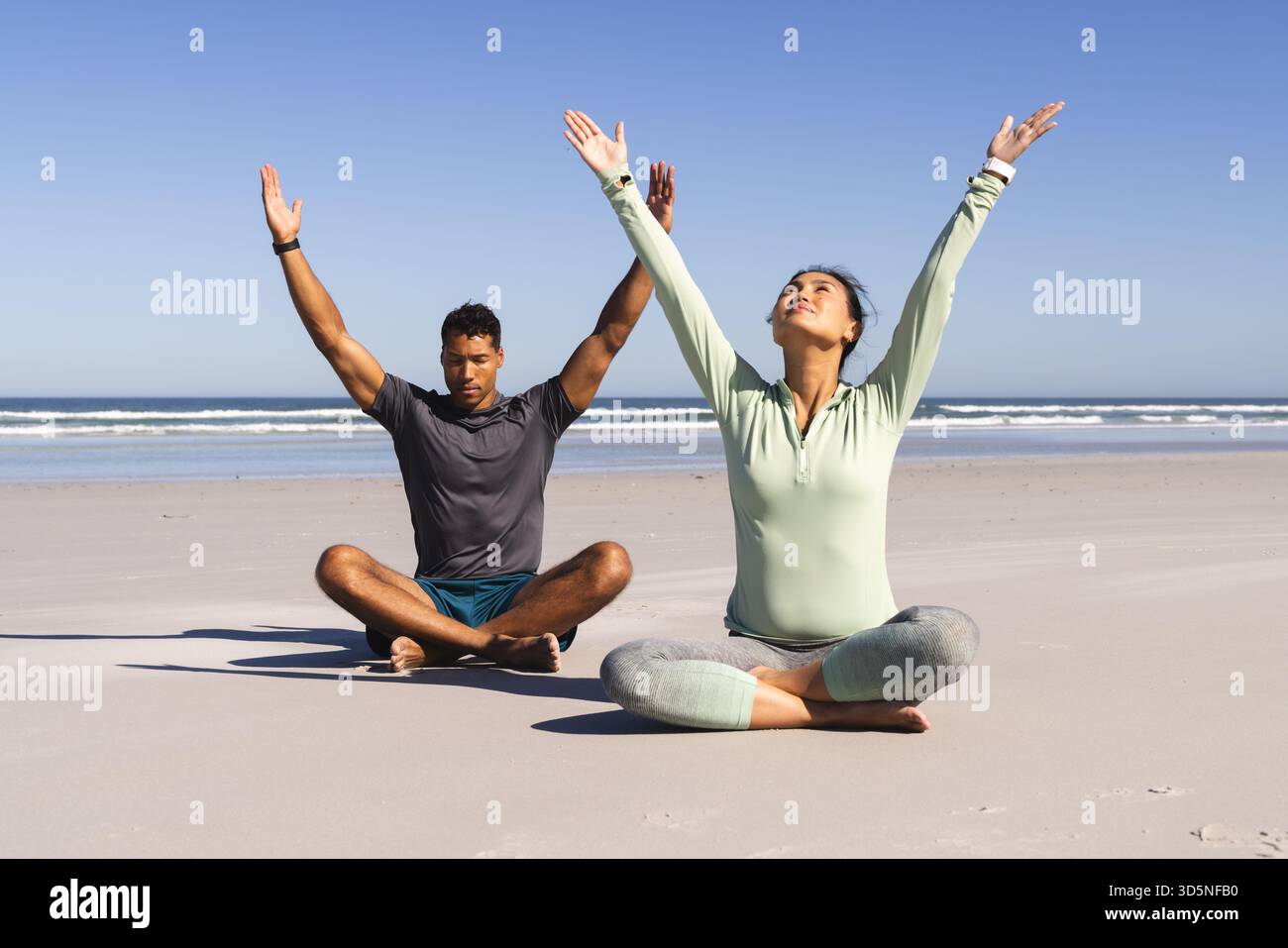 Coppia che pratica yoga sulla spiaggia, meditando con le braccia sollevate sotto il cielo limpido Foto Stock