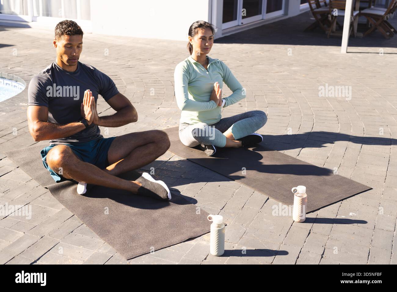 Coppie che praticano yoga all'aperto su tappeti, meditando con bottiglie d'acqua nelle vicinanze Foto Stock