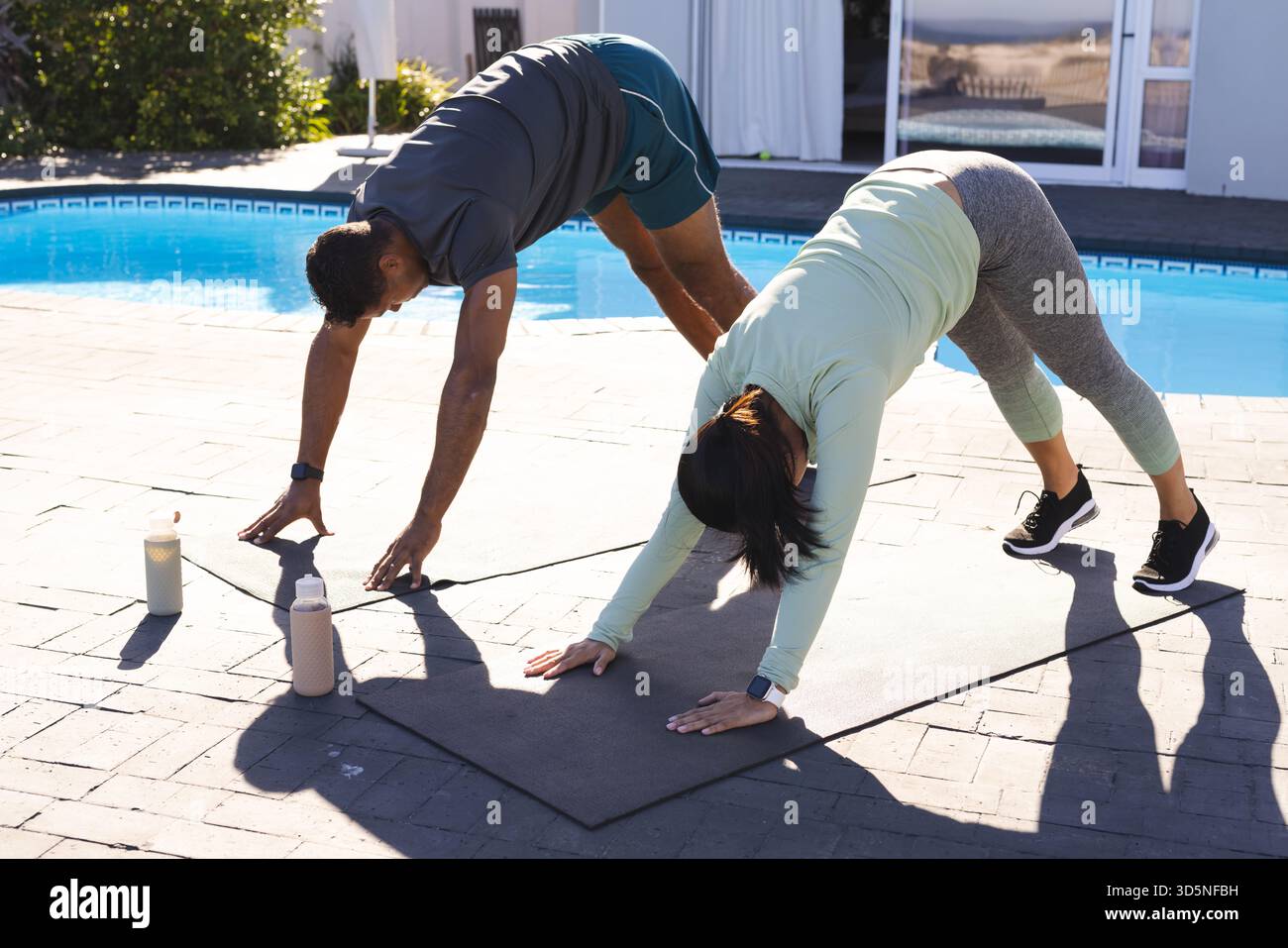 Coppie che praticano yoga all'aperto presso la piscina e si allungano sui tappetini alla luce del sole mattutino Foto Stock
