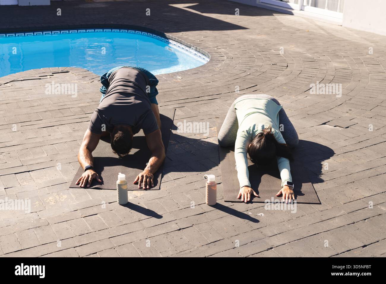 Coppie che praticano yoga in piscina, si allungano sui tappetini con bottiglie d'acqua nelle vicinanze Foto Stock