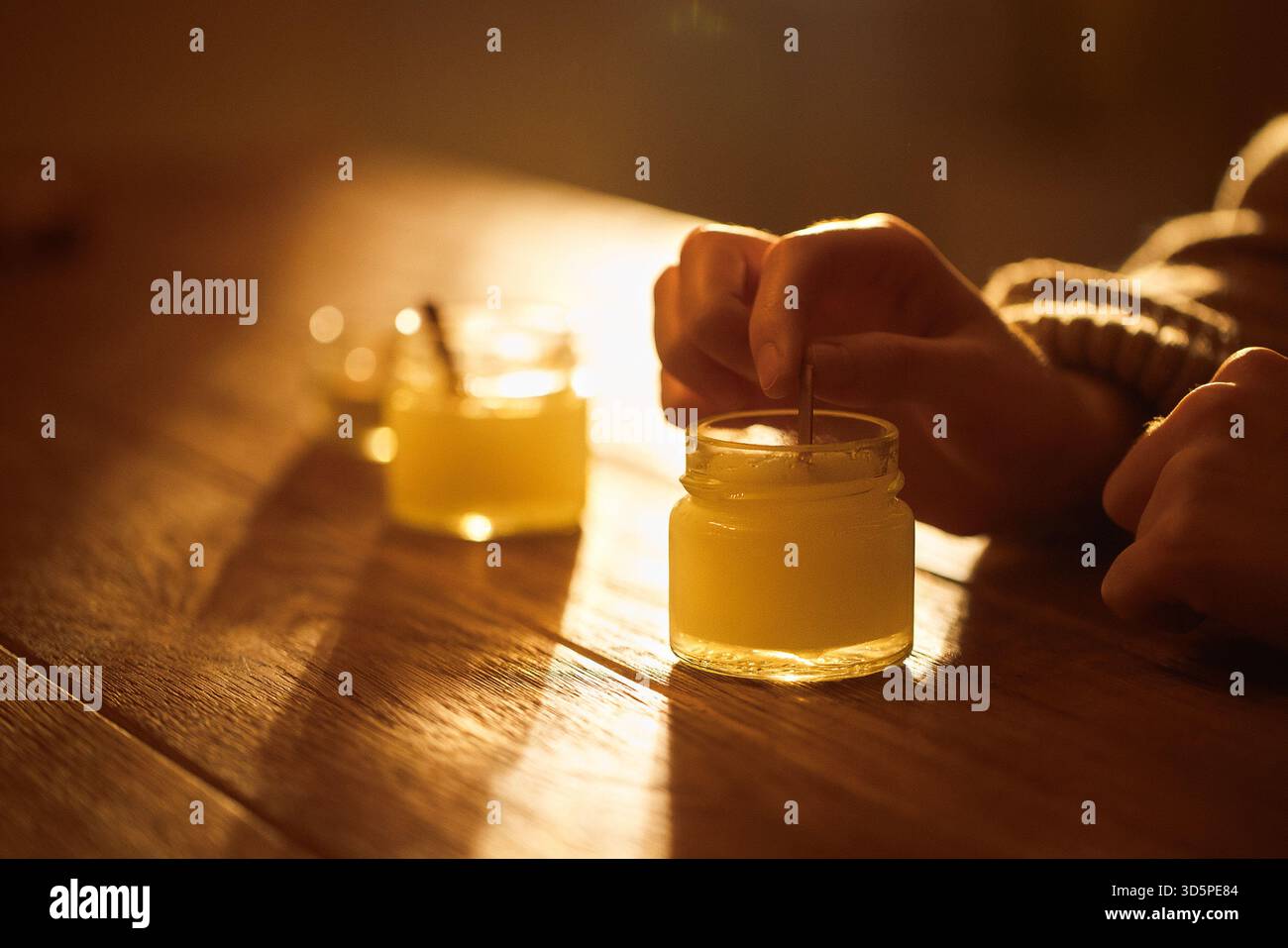 Le mani della persona mescolano delicatamente un vaso di candele su un tavolo in legno rustico, con luce solare calda che proietta ombre morbide, migliorando l'atmosfera accogliente del Foto Stock