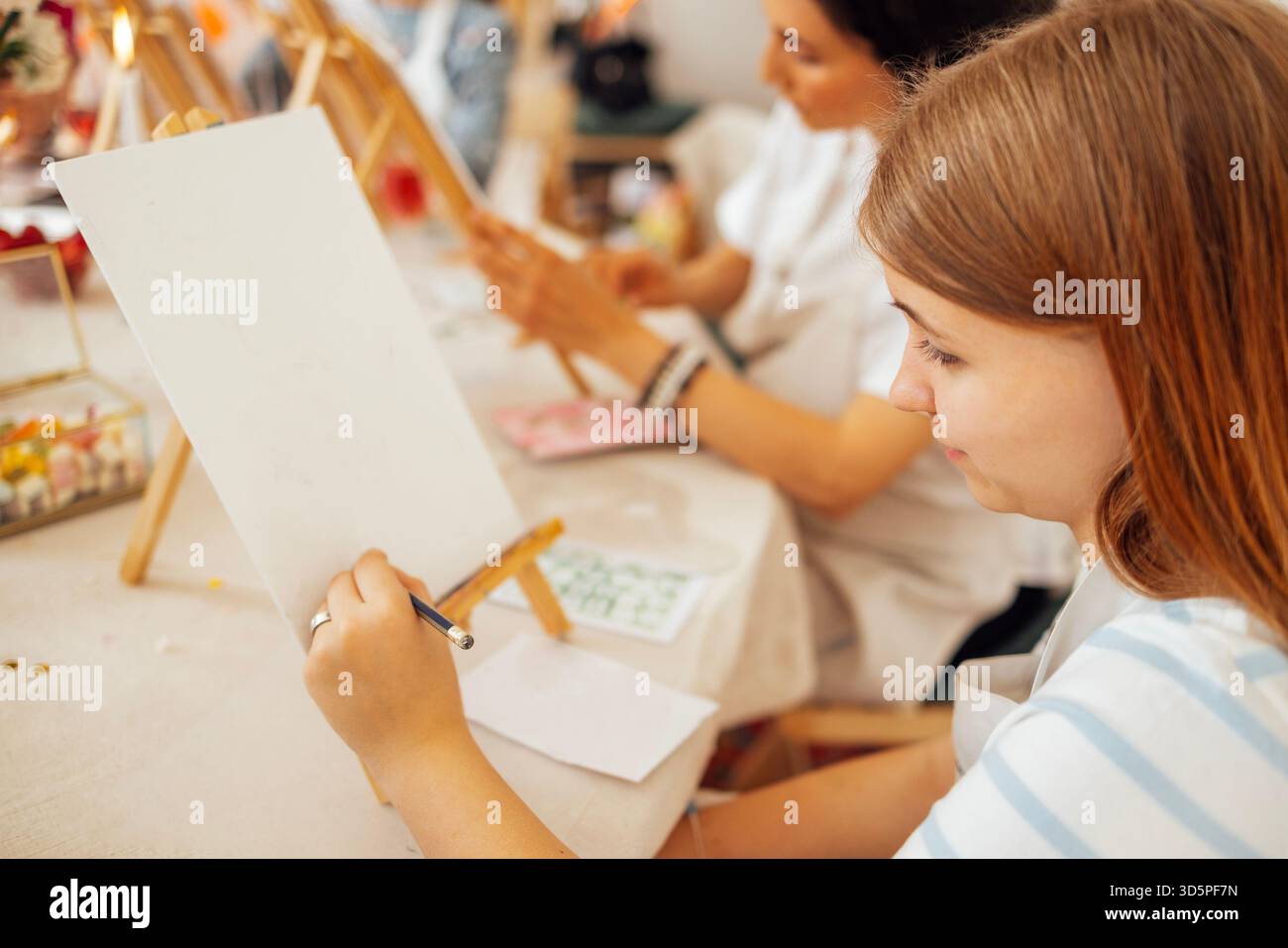 L'artista femminile si concentra sulla pittura su una tela vuota in un vivace laboratorio, con altri partecipanti impegnati in attività creative e expl artistiche Foto Stock