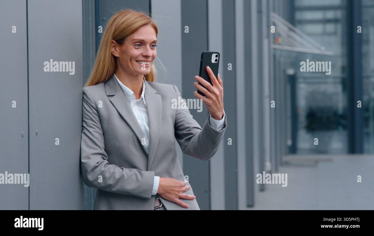 Donna d'affari caucasica di mezza età che parla videochiamata telefonica in città appoggiata al muro di un edificio di uffici donna d'affari adulta che chatta online Foto Stock