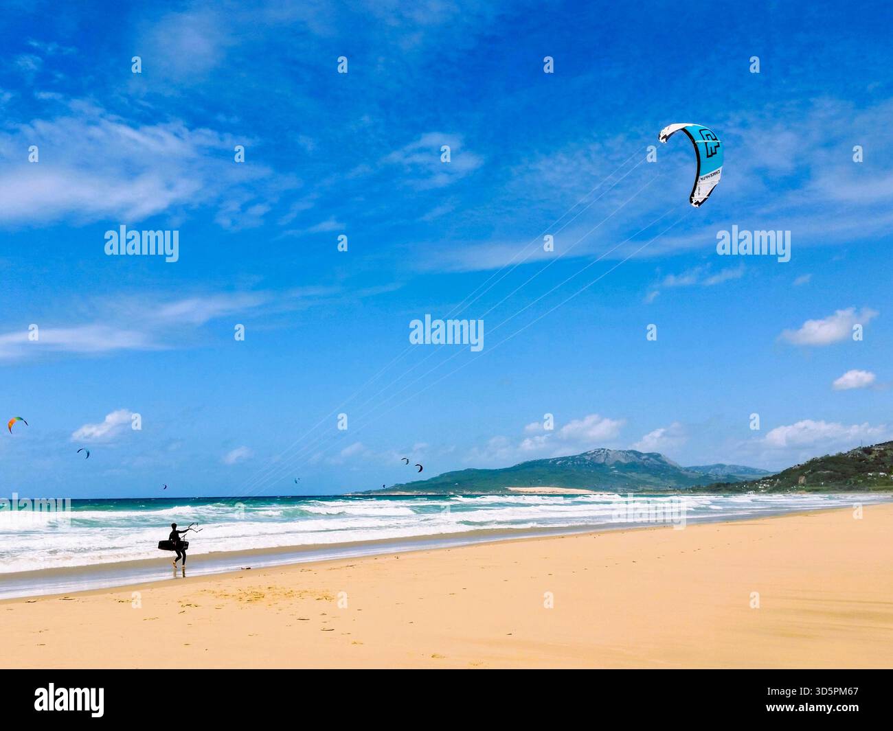 Un kitesurfer cammina lungo l'incontaminata spiaggia di sabbia bianca di Tarifa, Spagna. L'acqua blu turchese e le morbide onde bianche creano una vivace atmosfera costiera Foto Stock