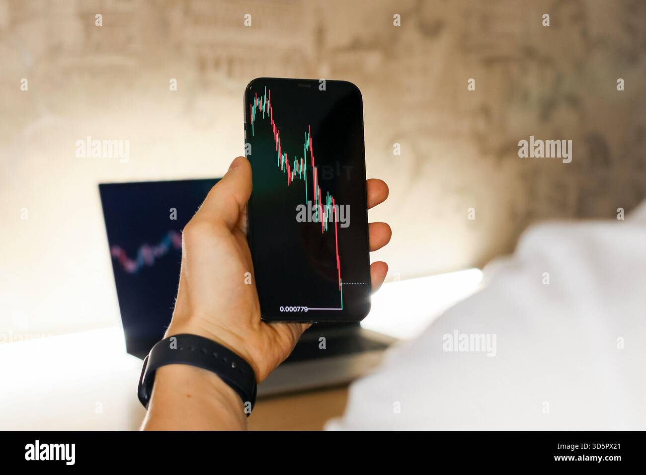 Primo piano della mano di una persona afferrando uno smartphone che mostra un grafico rosso del mercato azionario in declino, un laptop con un grafico simile visibile dietro una scrivania. Foto Stock