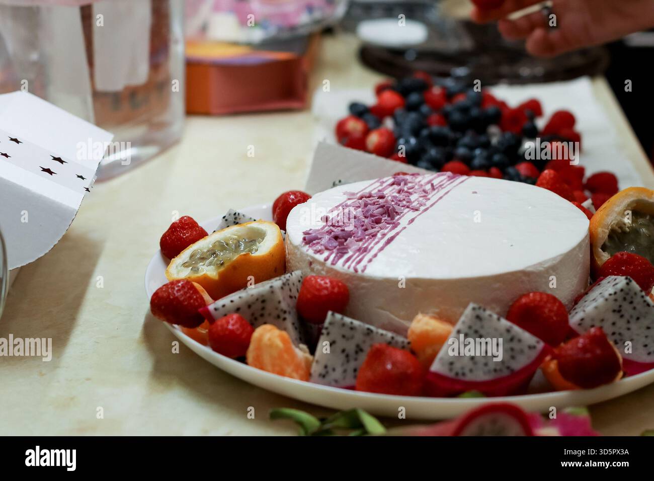 Vivace assortimento di frutta a fette e frutti di bosco disposti intorno a un dessert bianco rotondo con salsa viola su un piatto in cucina Foto Stock